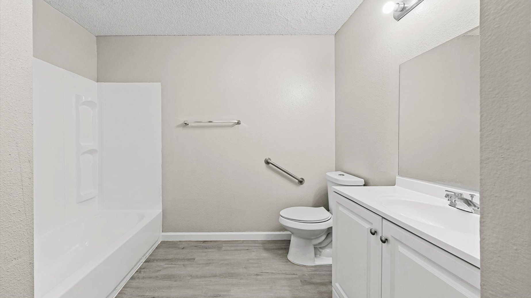 Bathroom interior with a white tub/shower, toilet, white vanity, and grab bar against gray walls.