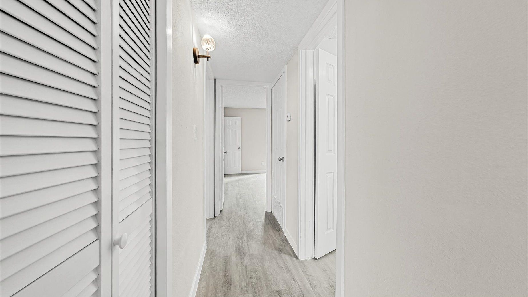Bright apartment hallway with white walls, doors, and a louvered closet on the left.