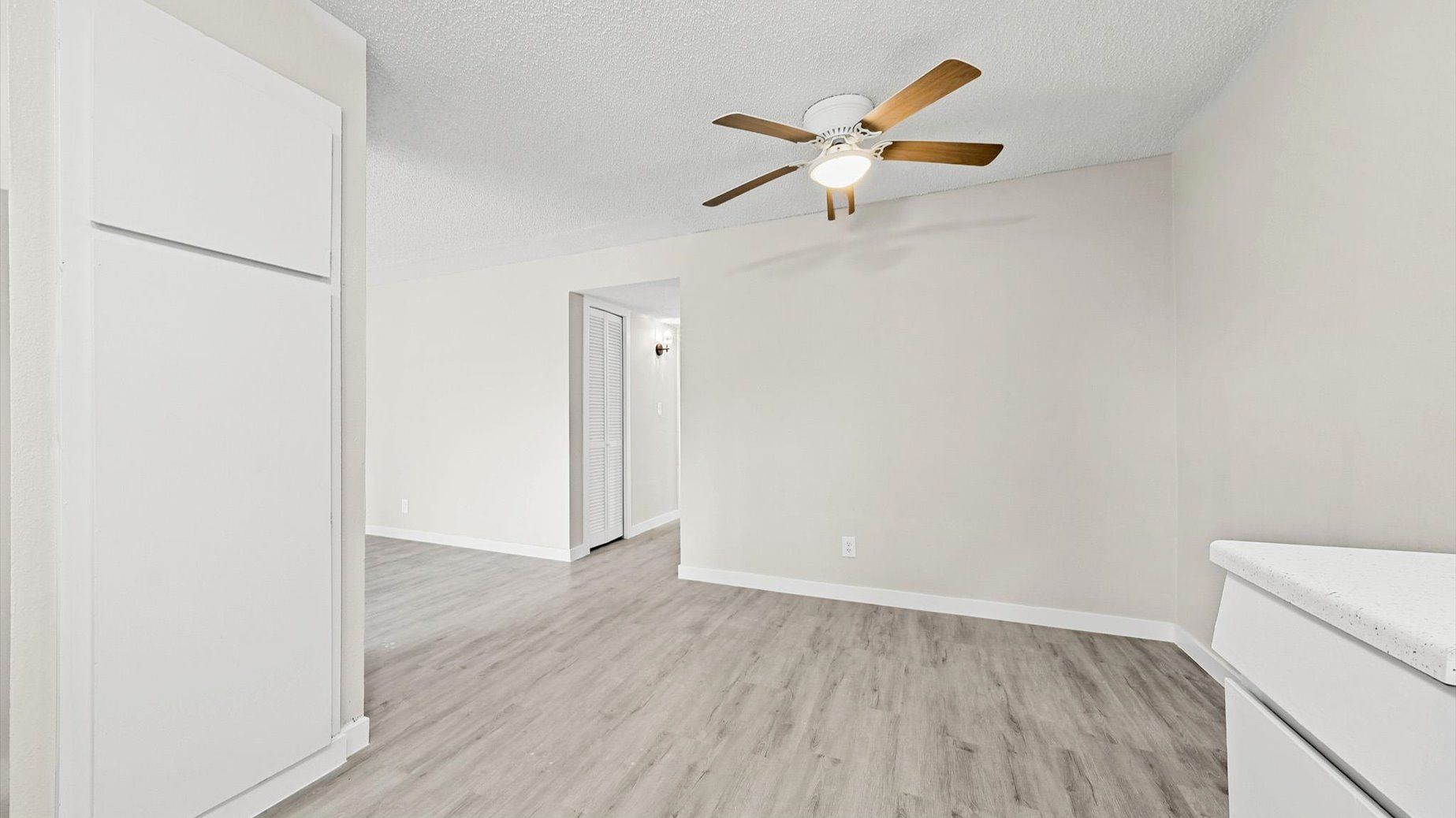 Empty apartment room with a ceiling fan, light, white closet doors, and light gray laminate flooring.