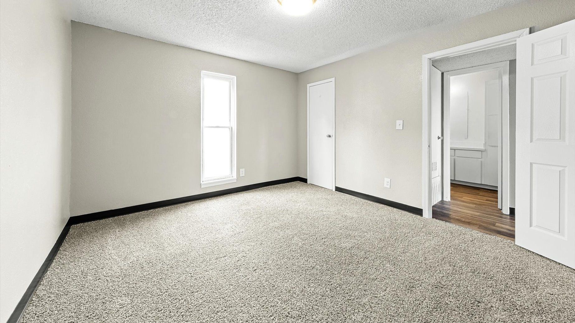 Empty carpeted room with a window, beige walls, and a doorway to a kitchen.