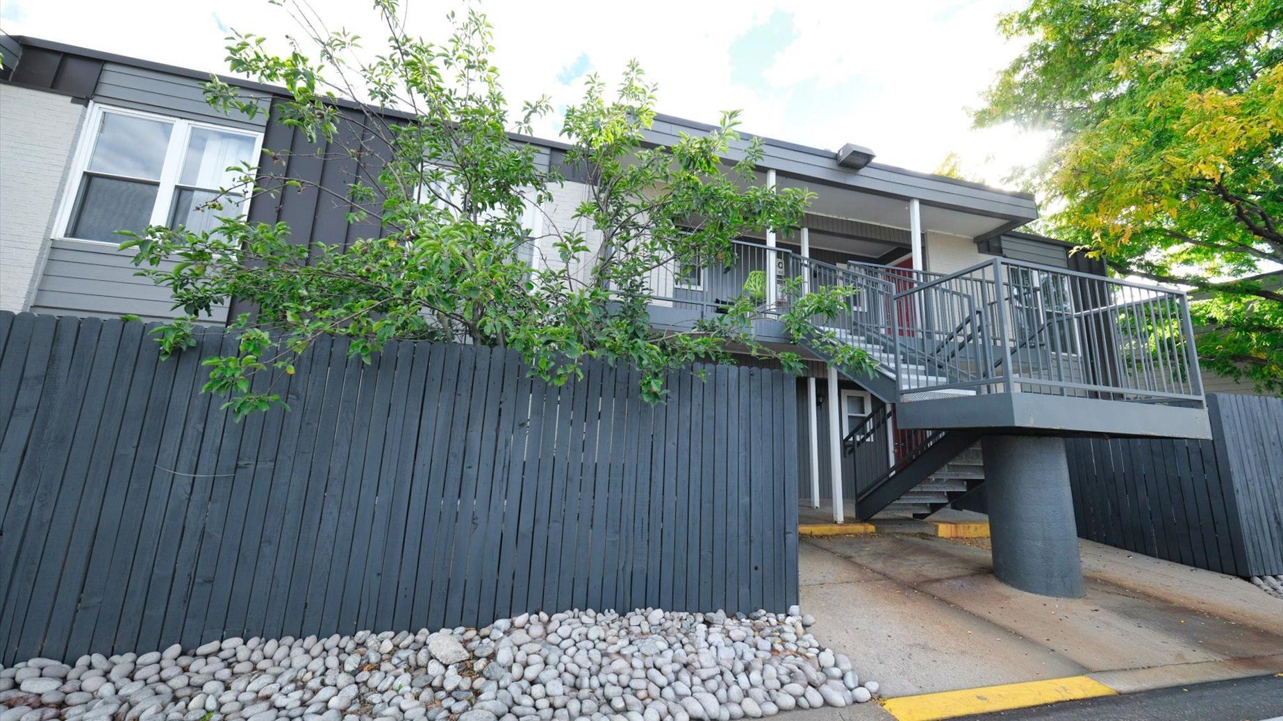 Exterior view of a multi-family building with a gray elevated deck, stairs, and fence.