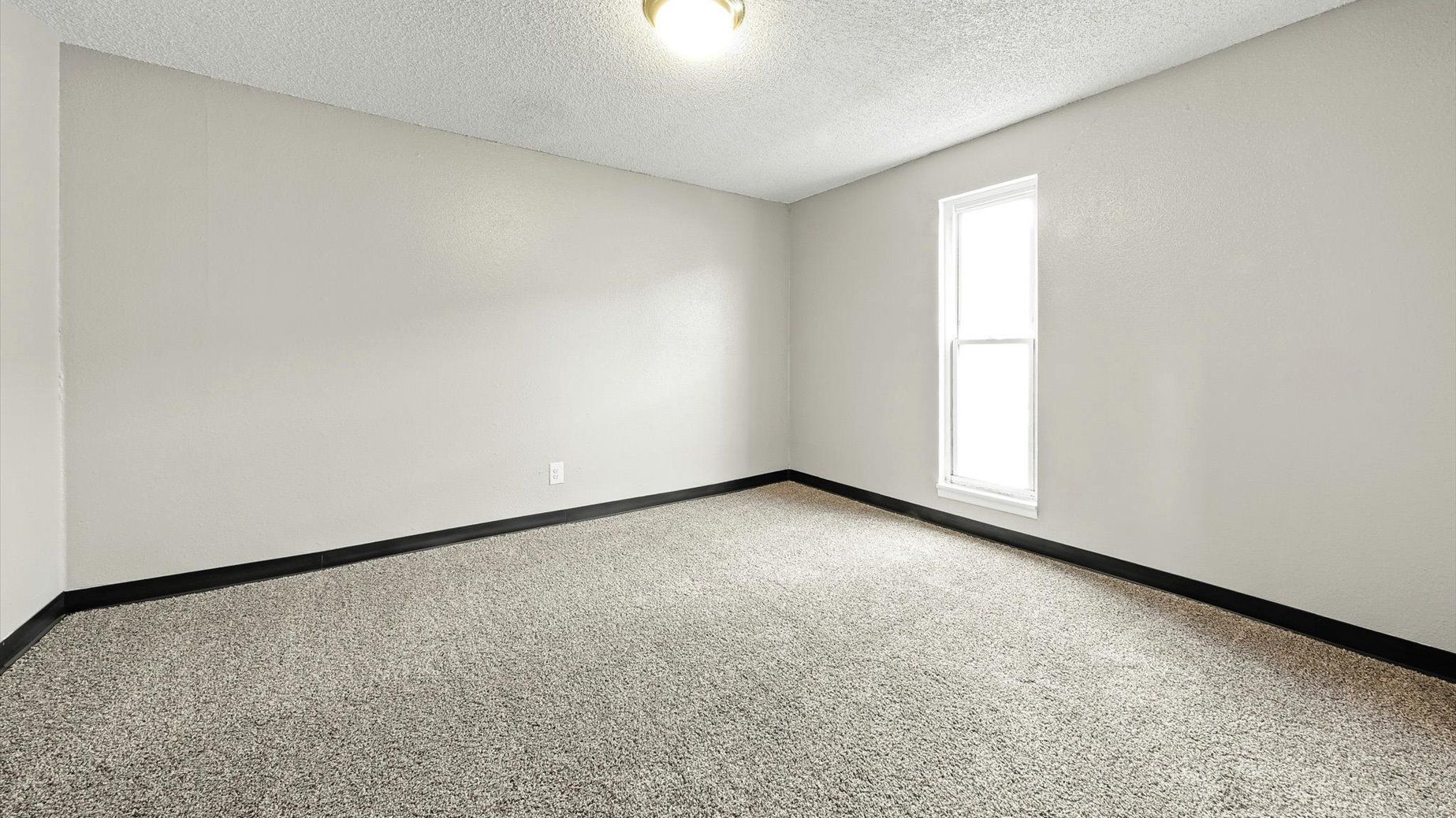 Empty carpeted room with a single window on the right and overhead ceiling light.