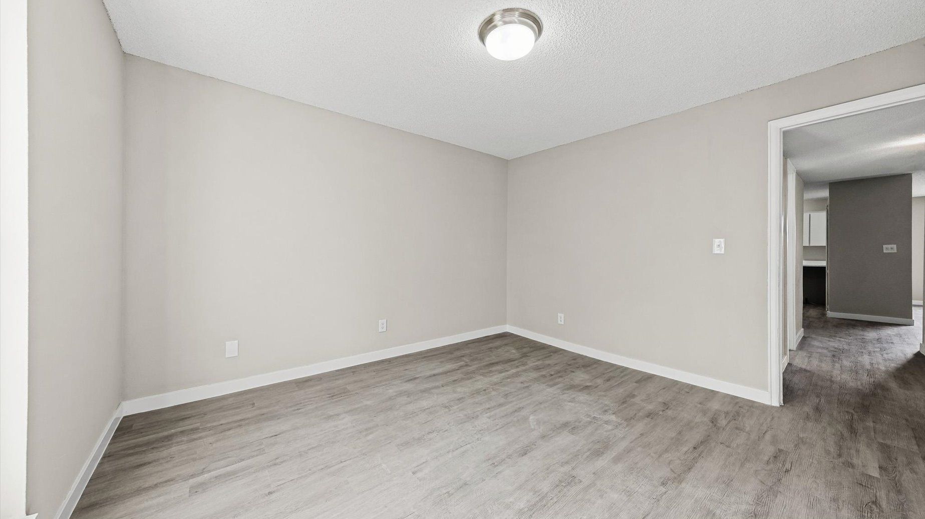 Empty beige-walled apartment room with gray flooring and a doorway to the kitchen.