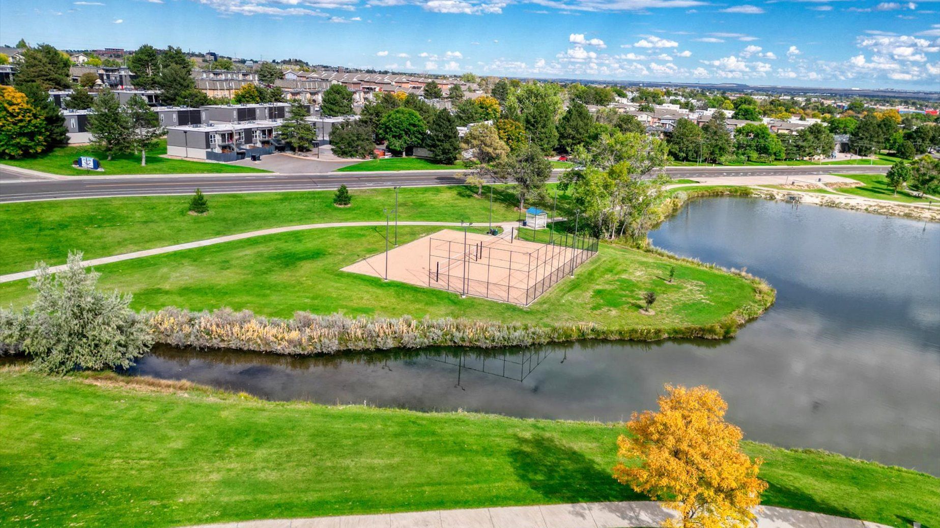Aerial view of a park-like community with a pond, green lawns, and a fenced sports court.