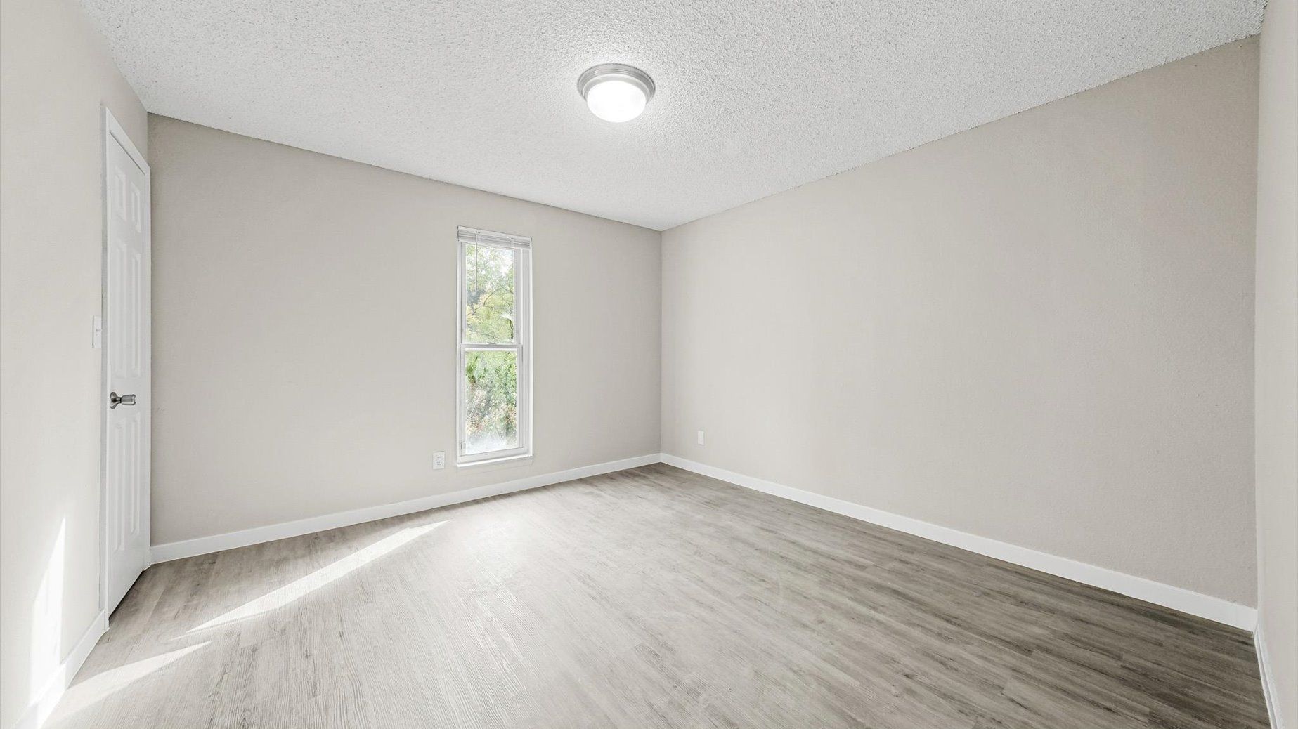 Empty beige room with a small window, door and light wood-like flooring.