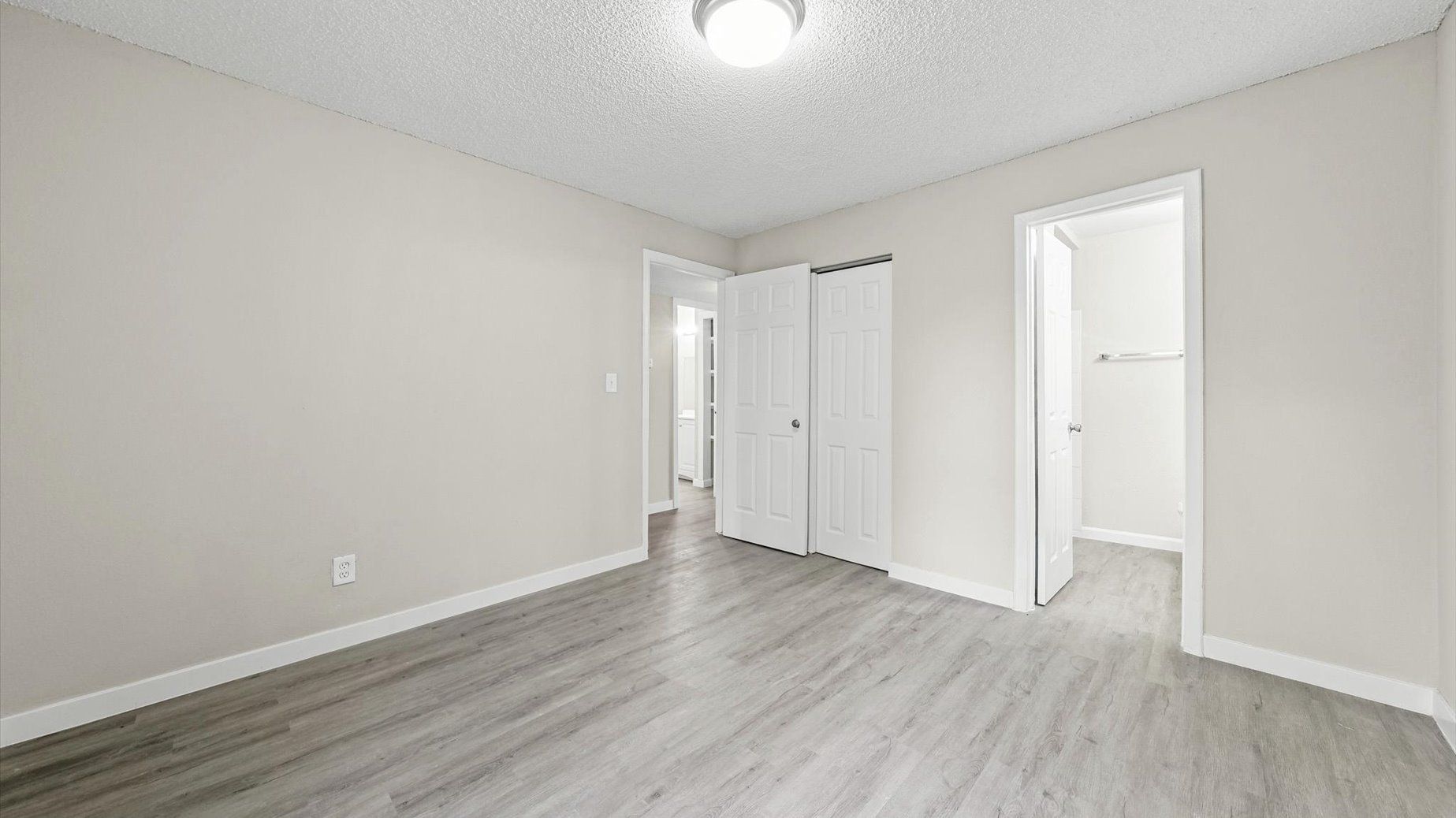 Empty bedroom in an apartment with beige walls, white doors, and light gray wood-look flooring.