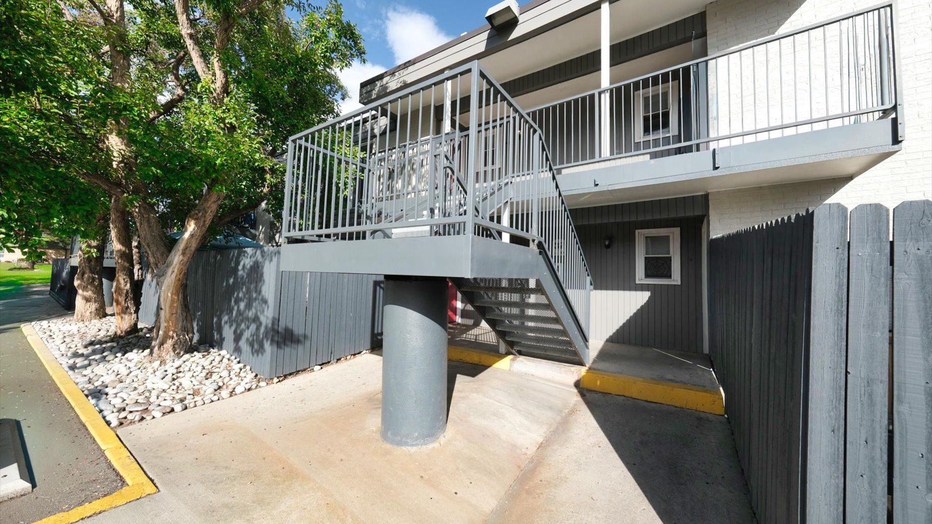 Exterior stairway with metal railing leading to upper-level apartments beside trees.