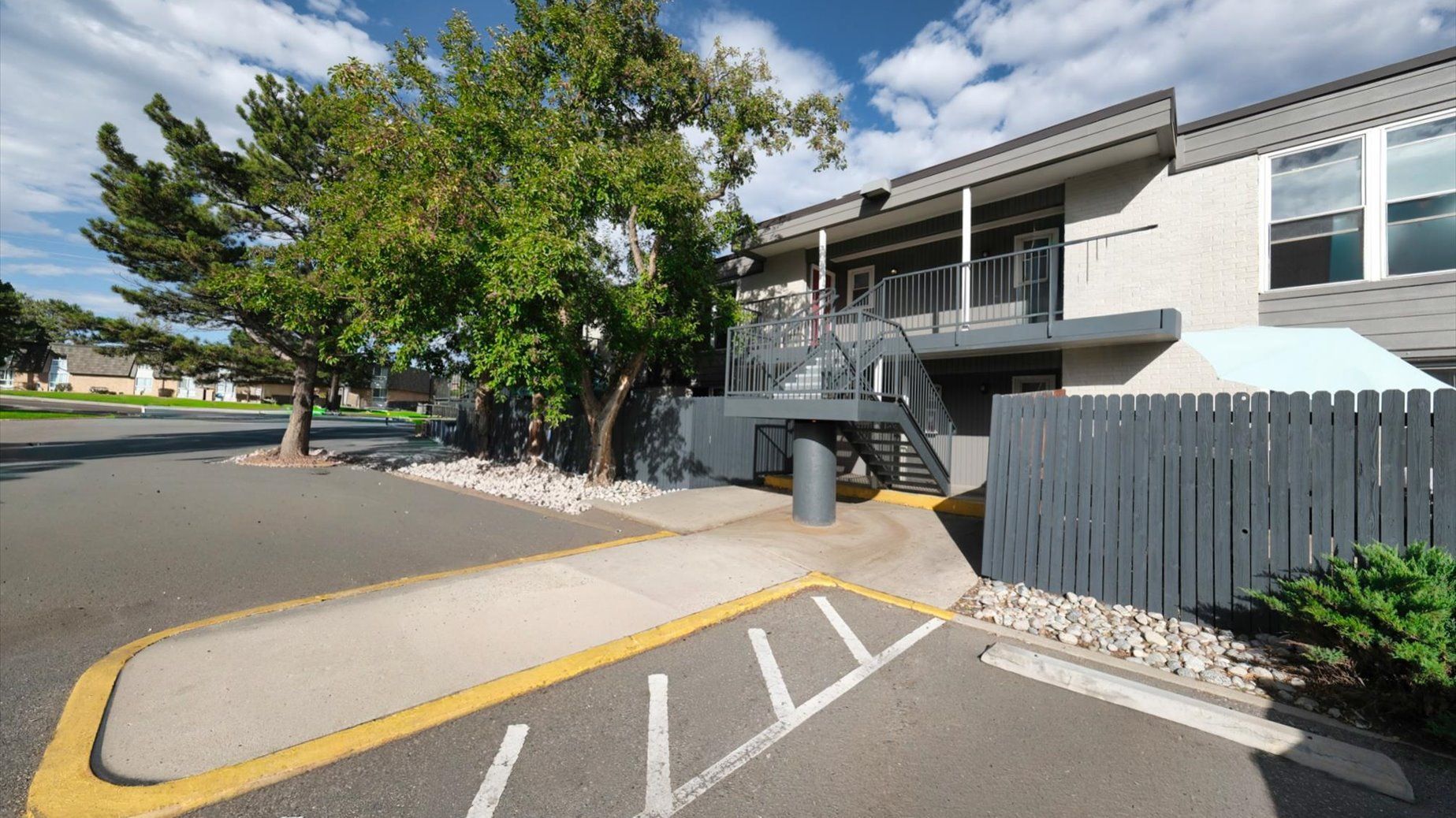 Exterior of an apartment building with stairs, gray railing, a wooden fence, trees, and a parking area.