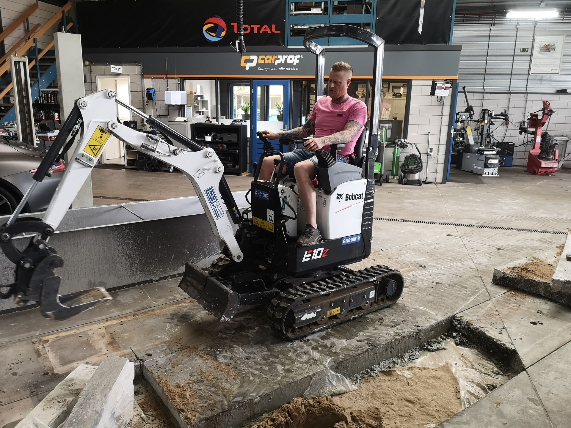 A man is sitting on a small excavator in a garage.