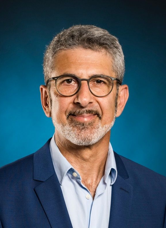 Mann mit Brille und grauem Bart in blauem Blazer lächelt vor blauem Hintergrund in die Kamera.