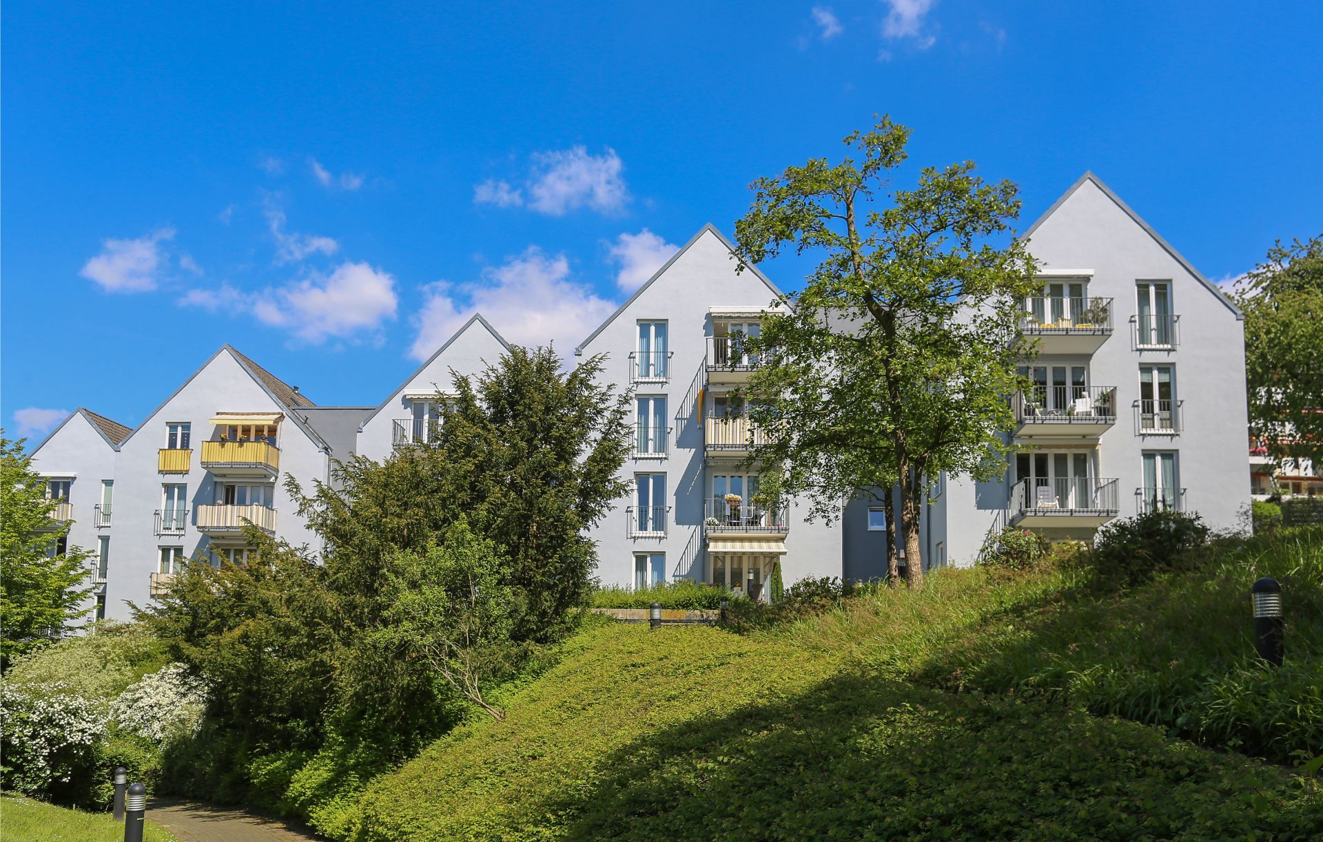 Graue Wohnhäuser mit schrägen Dächern und Balkonen, eingebettet in einen grasbewachsenen Hügel unter blauem Himmel.