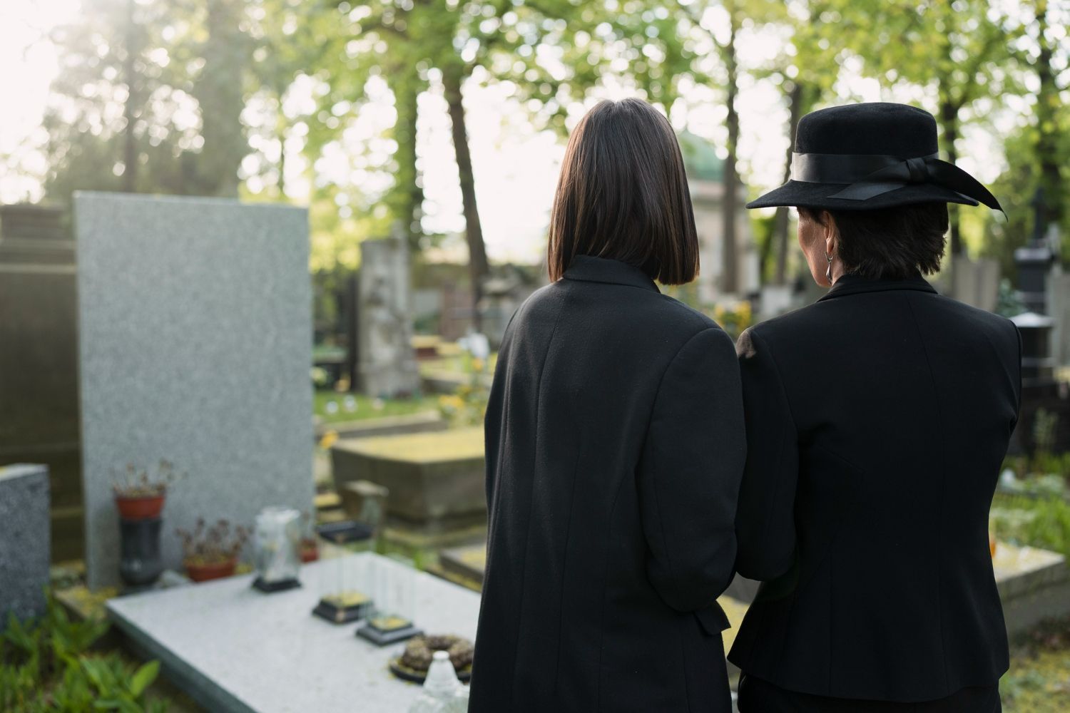 Two people in black clothing standing beside a cemetery grave marker outdoors