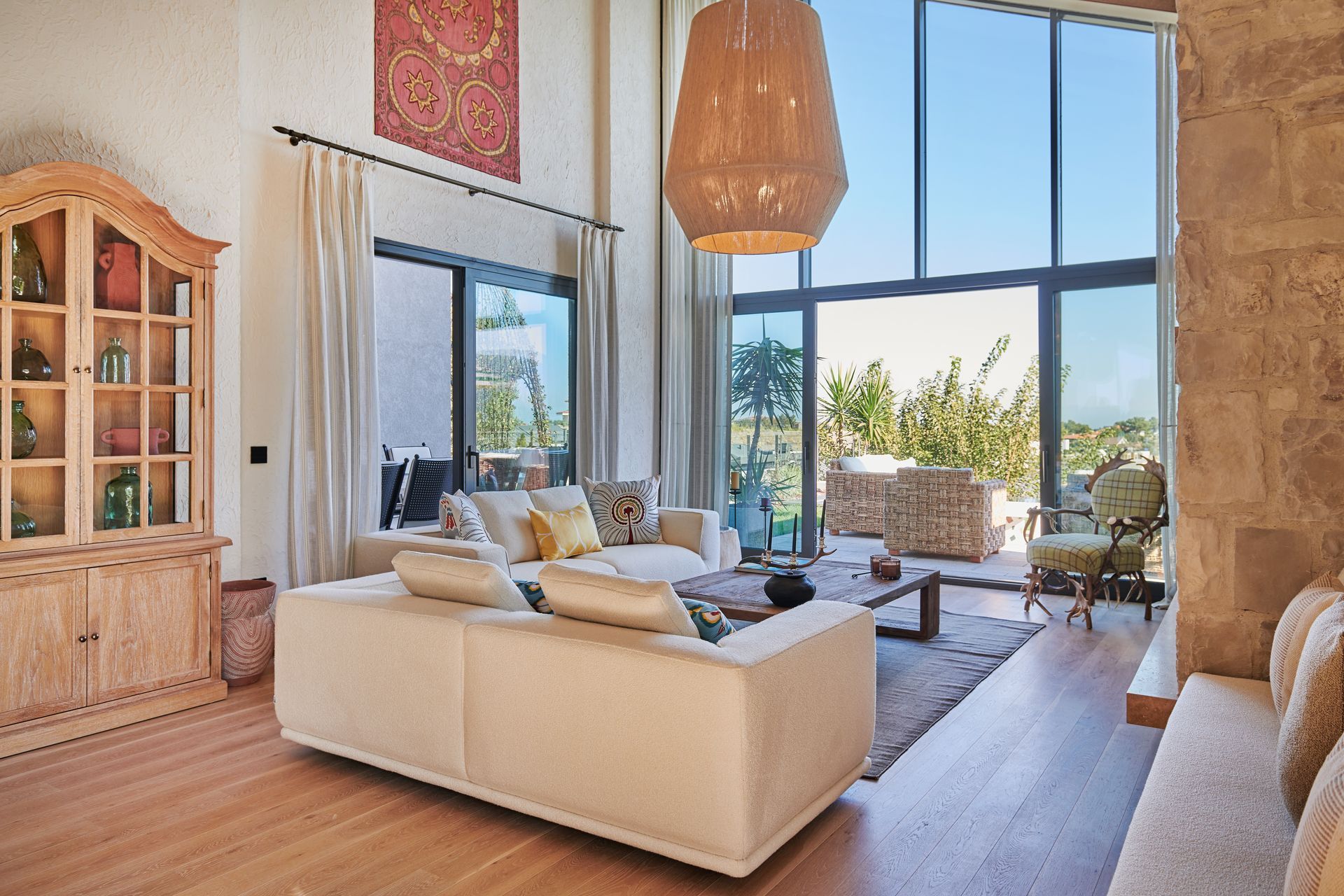 Living room with a high ceiling, large windows, two cream sofas, and a wooden display cabinet.