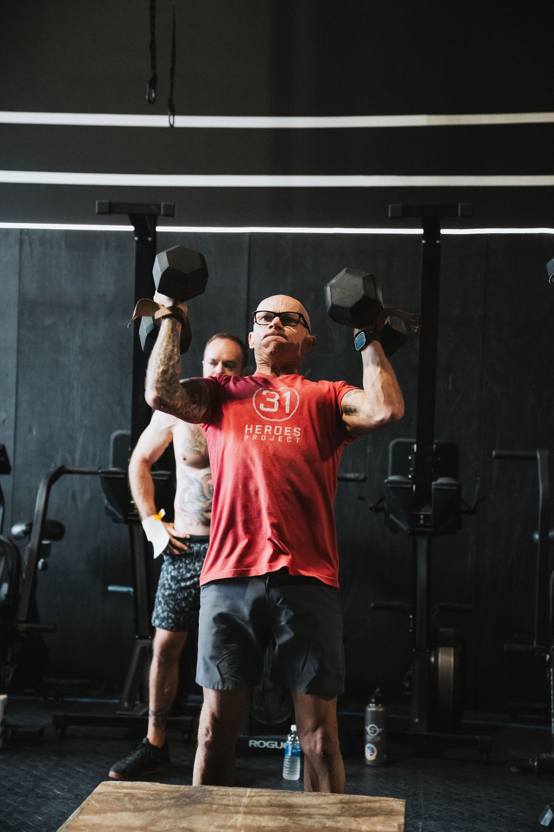 Man in red shirt lifting dumbbells in a gym, another man watches.