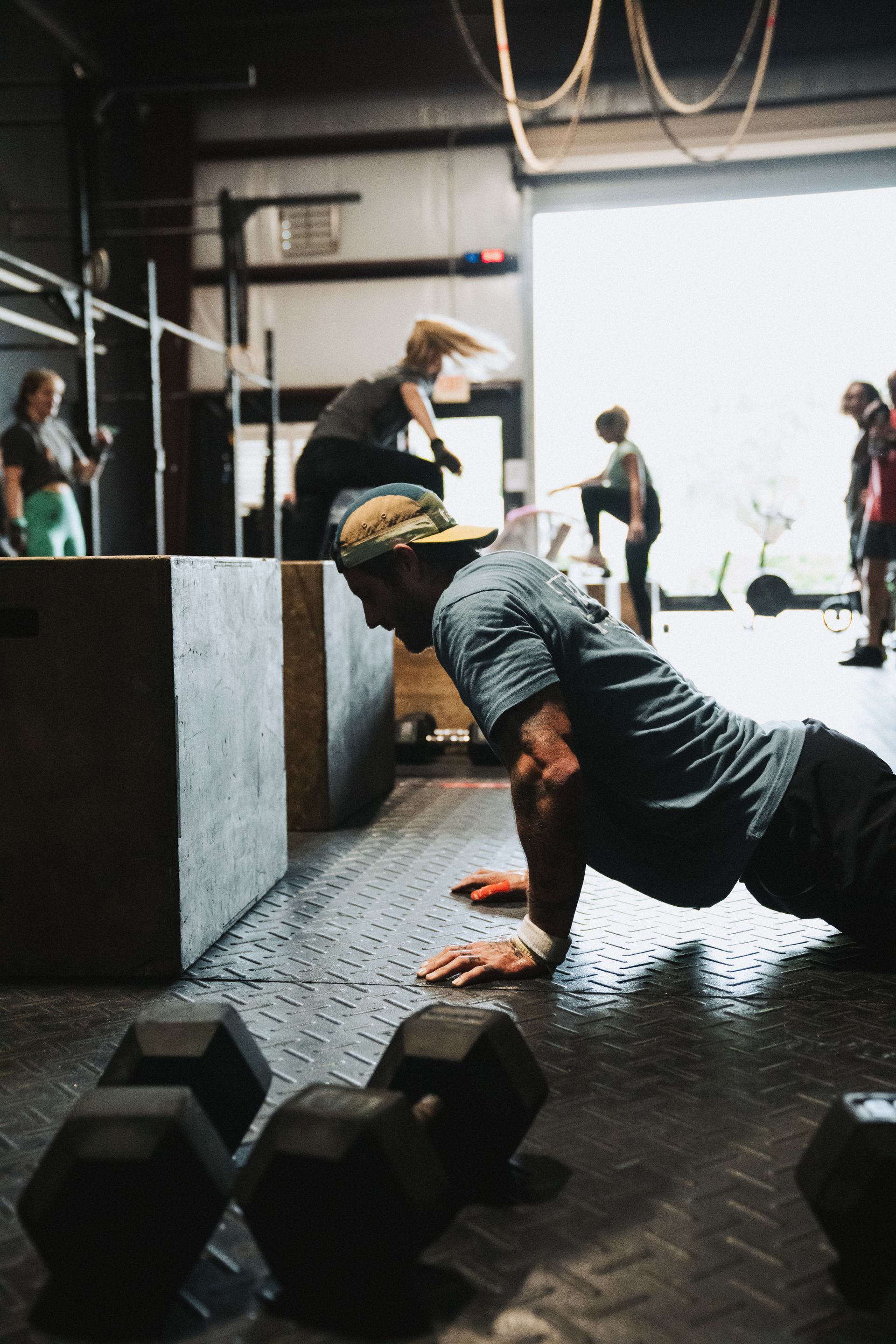 Man doing push-ups in gym, while a woman jumps over a box. Others exercise in the background.