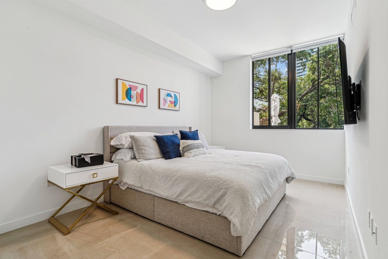 Furnished bedroom in a modern apartment featuring a bed, nightstand, large window, and wall-mounted TV.