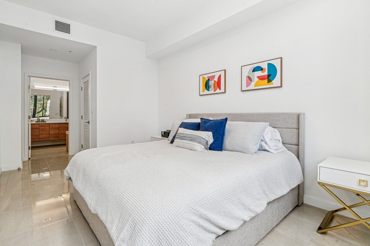 Bedroom with a large bed, white bedding, two colorful abstract art prints, and a white nightstand.