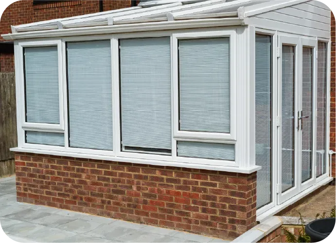 A white conservatory with blinds on the windows and a brick wall.