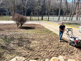 Man mowing muddy grass near a river, yellow caution tape, bare trees in the background.