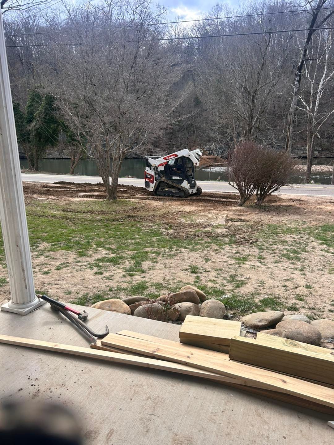 White and red motorcycle on a road by trees and a stream. Lawn and porch in foreground with tools.