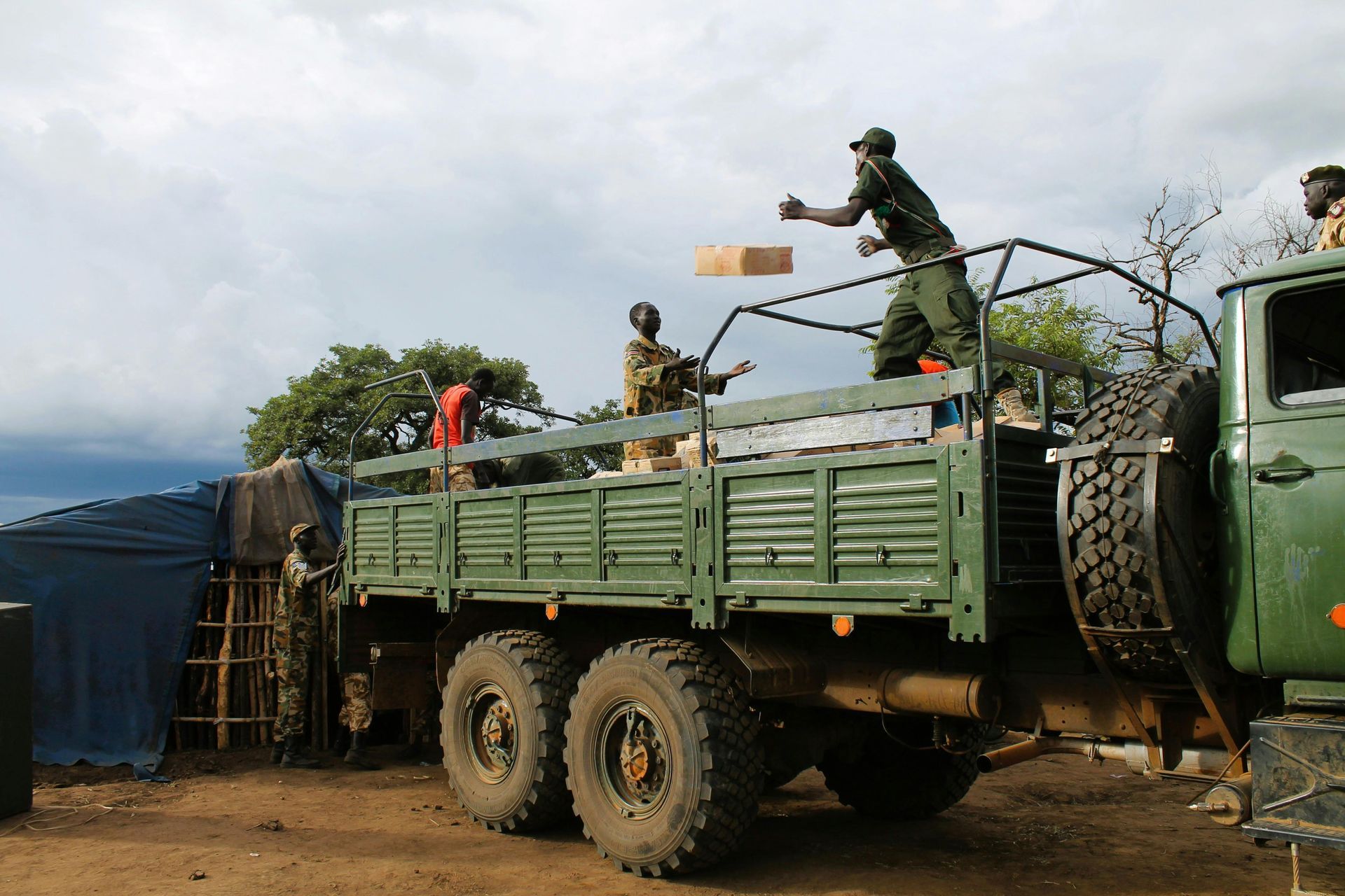 Soldiers unloading boxes from a large green truck outdoors under a cloudy sky.