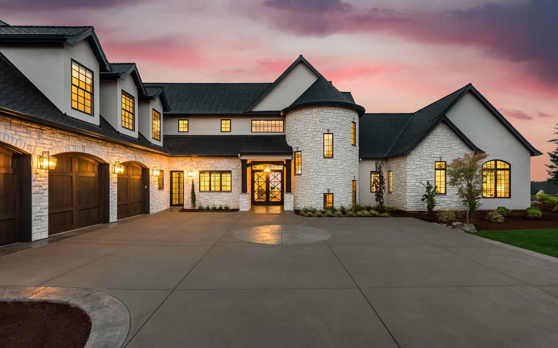 Large modern stone house at dusk with lit windows, three-car garage, and wide driveway under a pink sky