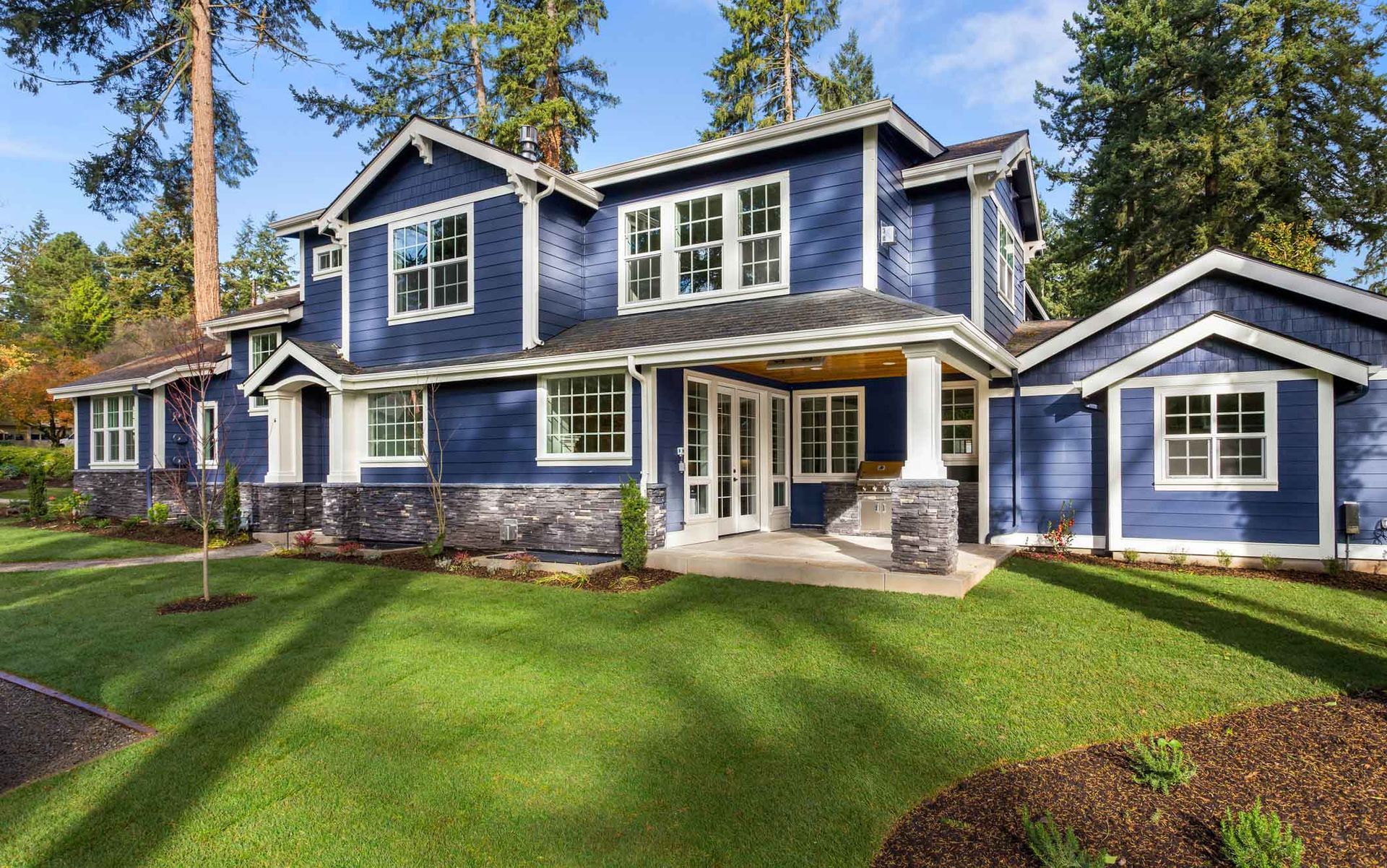 Blue two-story house with white trim and a front porch on a green lawn surrounded by tall trees