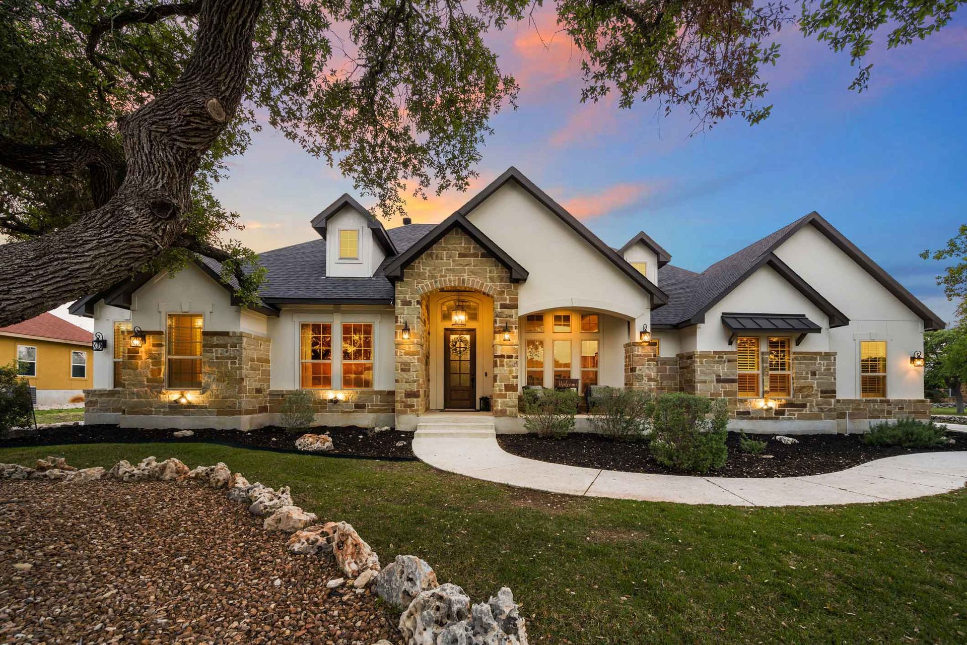 Elegant stone-and-stucco house at dusk with lit windows, arched entry, and a curved front walkway