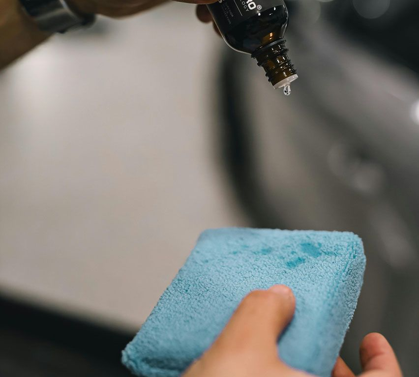 Person dispensing liquid from a bottle onto a blue microfiber cloth near a car.