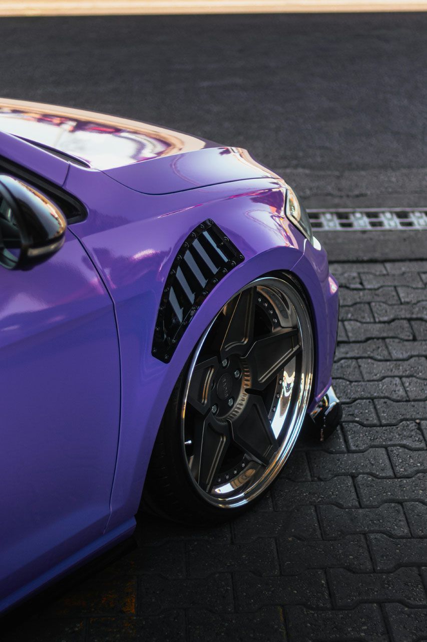 Purple car fender with chrome wheels, black vent, and low stance on a brick surface.