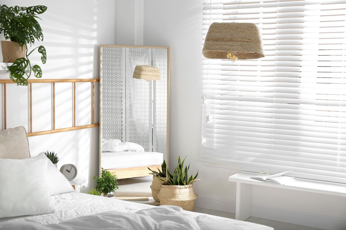 A bedroom with a bed , mirror , plants and blinds.