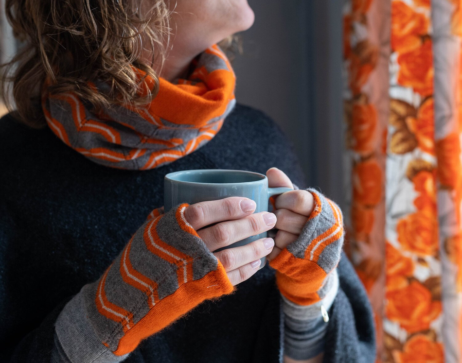 Woman holding a coffee mug, wearing fingerless gloves with a brightly coloured zigzag print and matching neck warmer.
