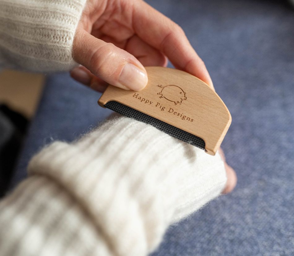 A hand using a small wooden fabric comb, with Happy Pig Designs logo on, to de-pill cashmere wrist warmers.