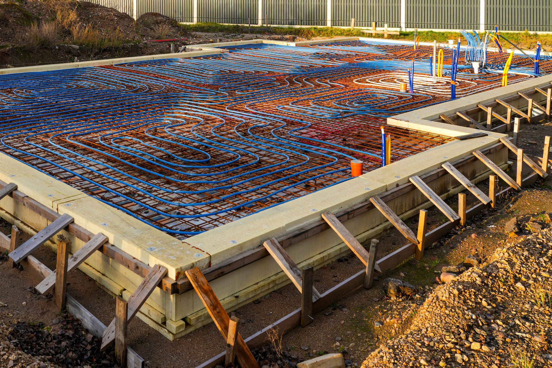 Construction site with concrete foundation frame, blue and orange pipes, and reinforcing rebar.