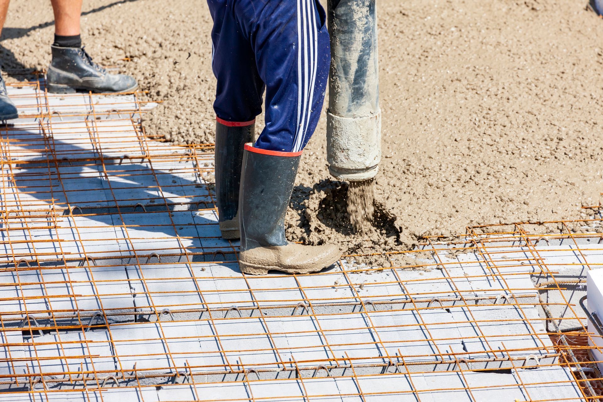 Worker pouring concrete over a steel rebar grid during a foundation construction process.