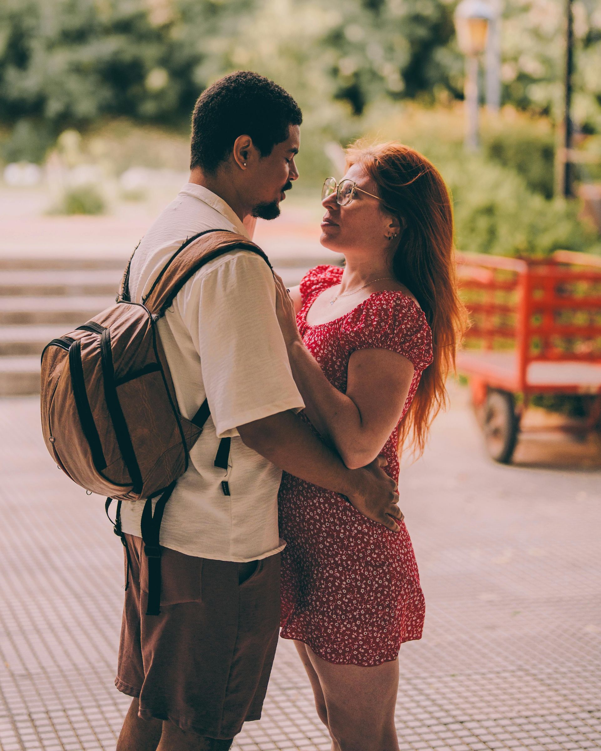 Two people embracing outdoors, one with a backpack, near a red cart in the background.
