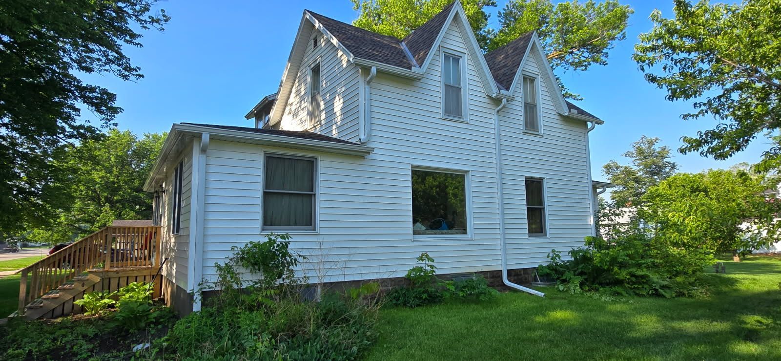 White two-story house with green shutters and a wooden ramp, surrounded by trees and grass under a sunny sky