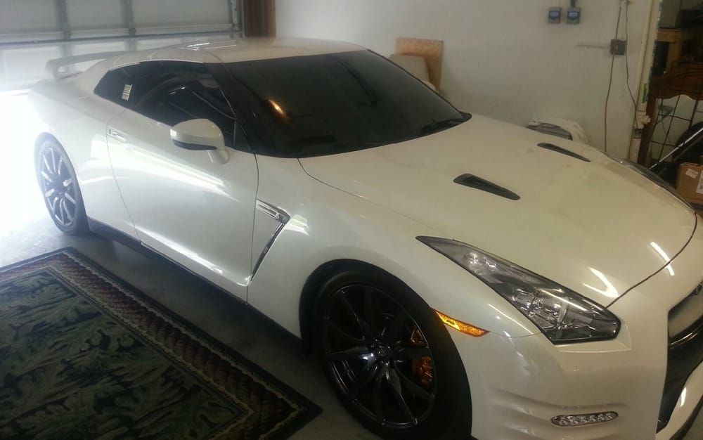 White Nissan GT-R sports car parked in a garage. Black wheels, tinted windows, and a dark rug.