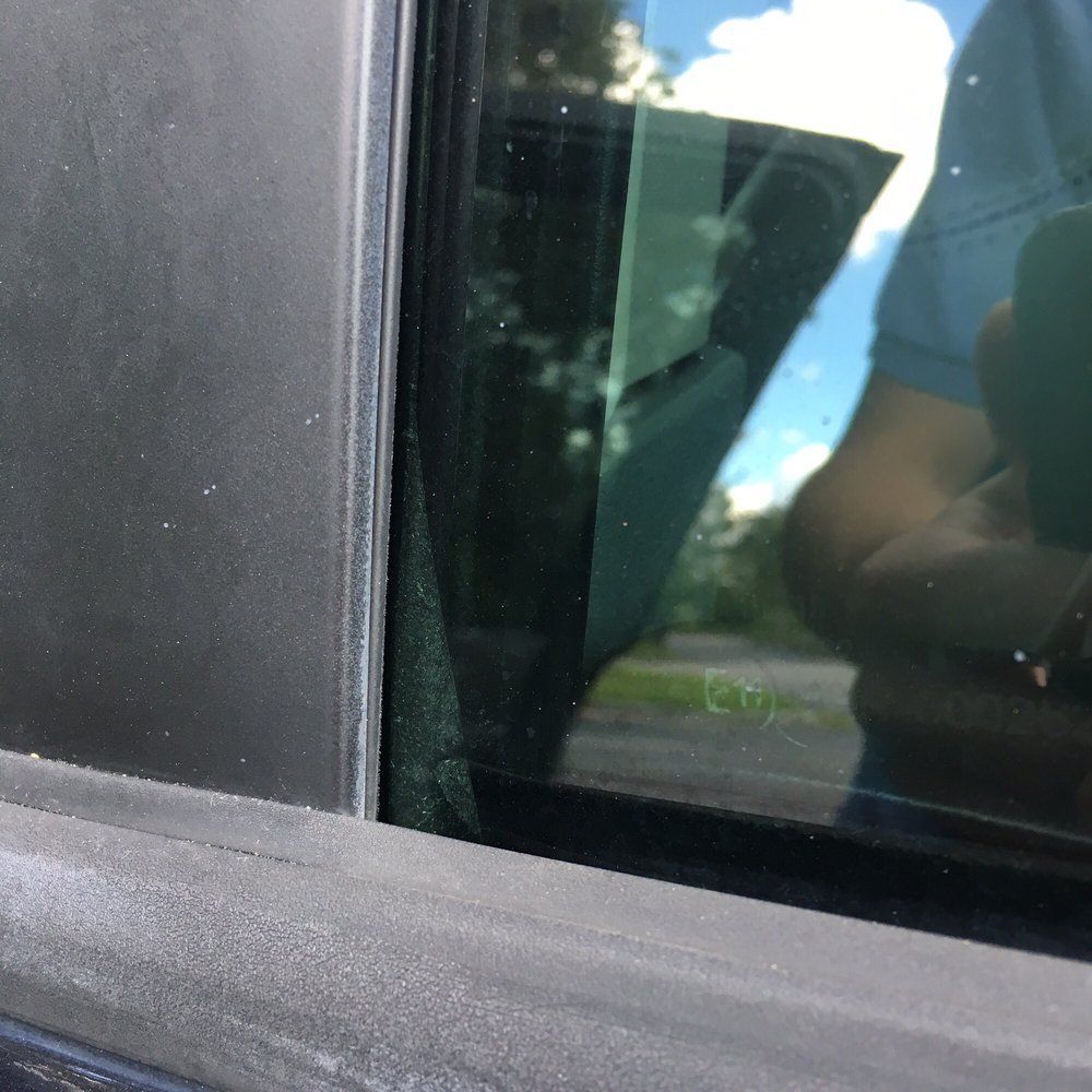 Close-up of a car window. Exterior trim is dark gray. Green tinted window reflects a person and sky.