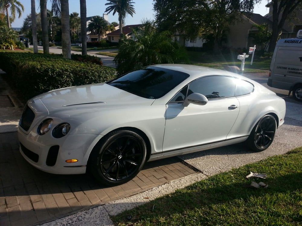 White Bentley coupe with black rims parked on a driveway in a sunny outdoor setting.