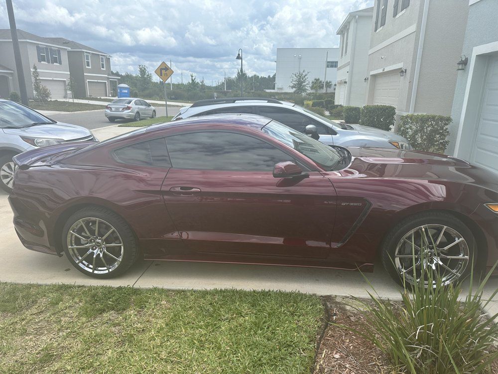 Burgundy Ford Mustang parked on a residential driveway, other cars and houses in background.