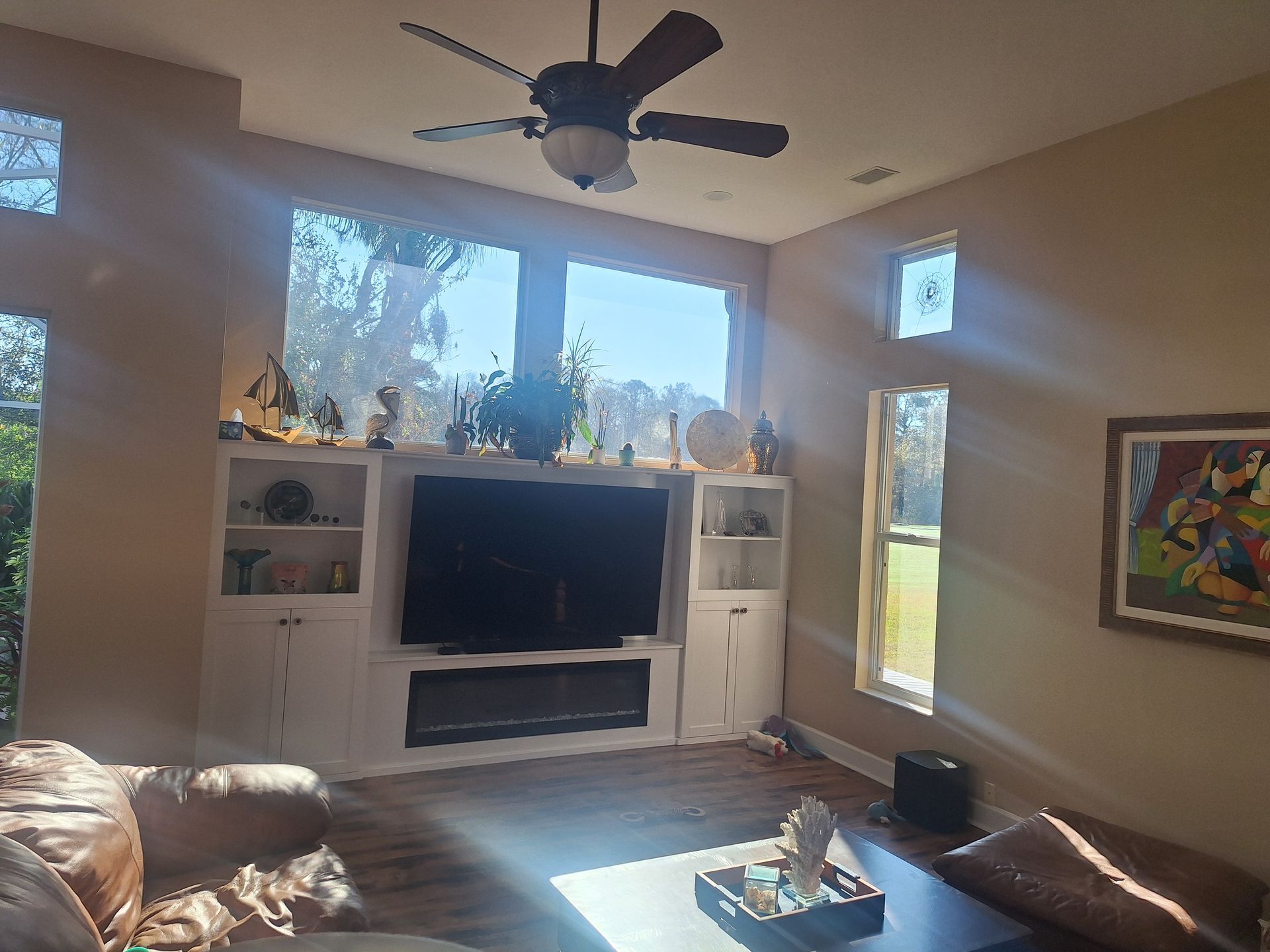 Living room with built-in media center, large windows, ceiling fan, and brown sofa. Bright sunlight.