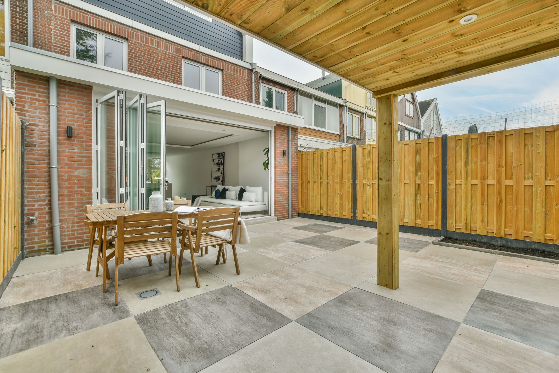 Terrasse avec table et chaises en bois, attenante à une maison aux portes ouvertes. Clôture en bois en arrière-plan.