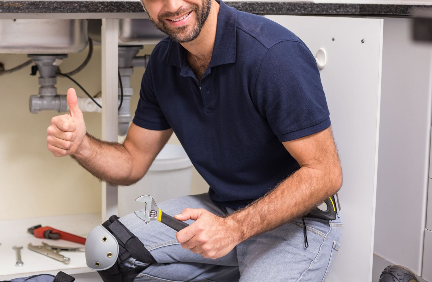 Plombier en chemise bleue, agenouillé, pouce levé. Travaillant sous un évier, une clé à molette à la main, il sourit.