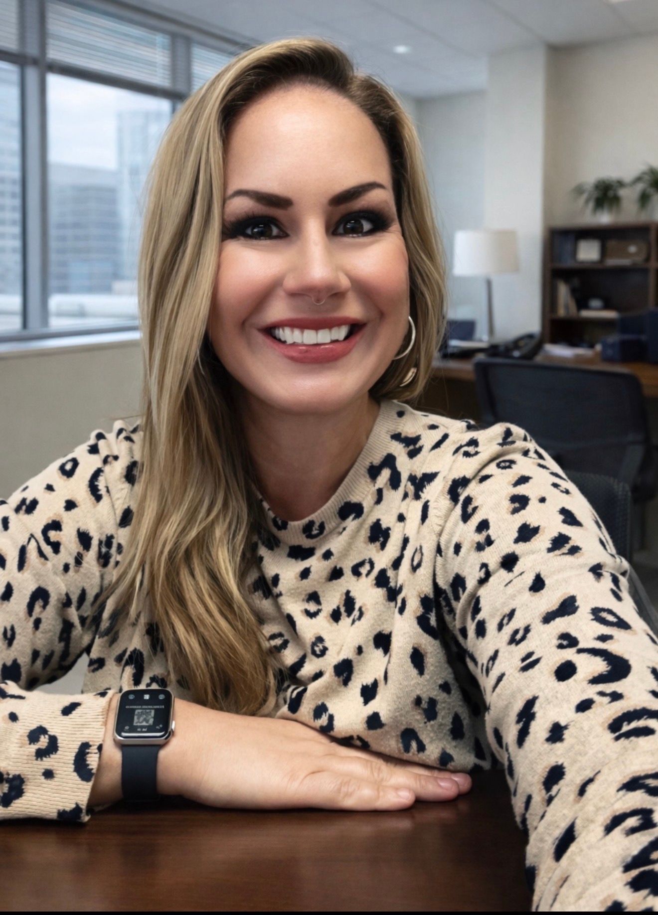 A smiling person with long blonde hair wearing a tan leopard-print sweater and a digital watch, posing in an office.