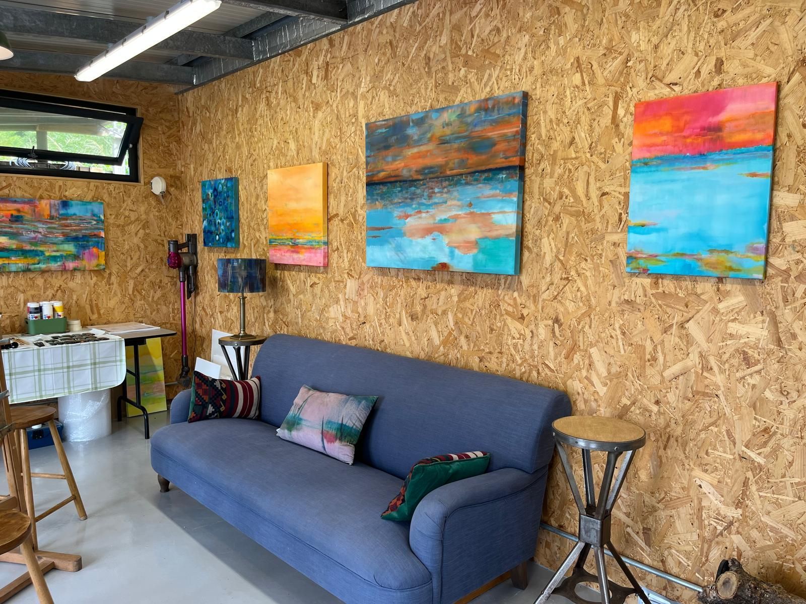 Cosy art studio with abstract paintings on wood panel walls, blue couch, and small table with acyrlic paints on it.