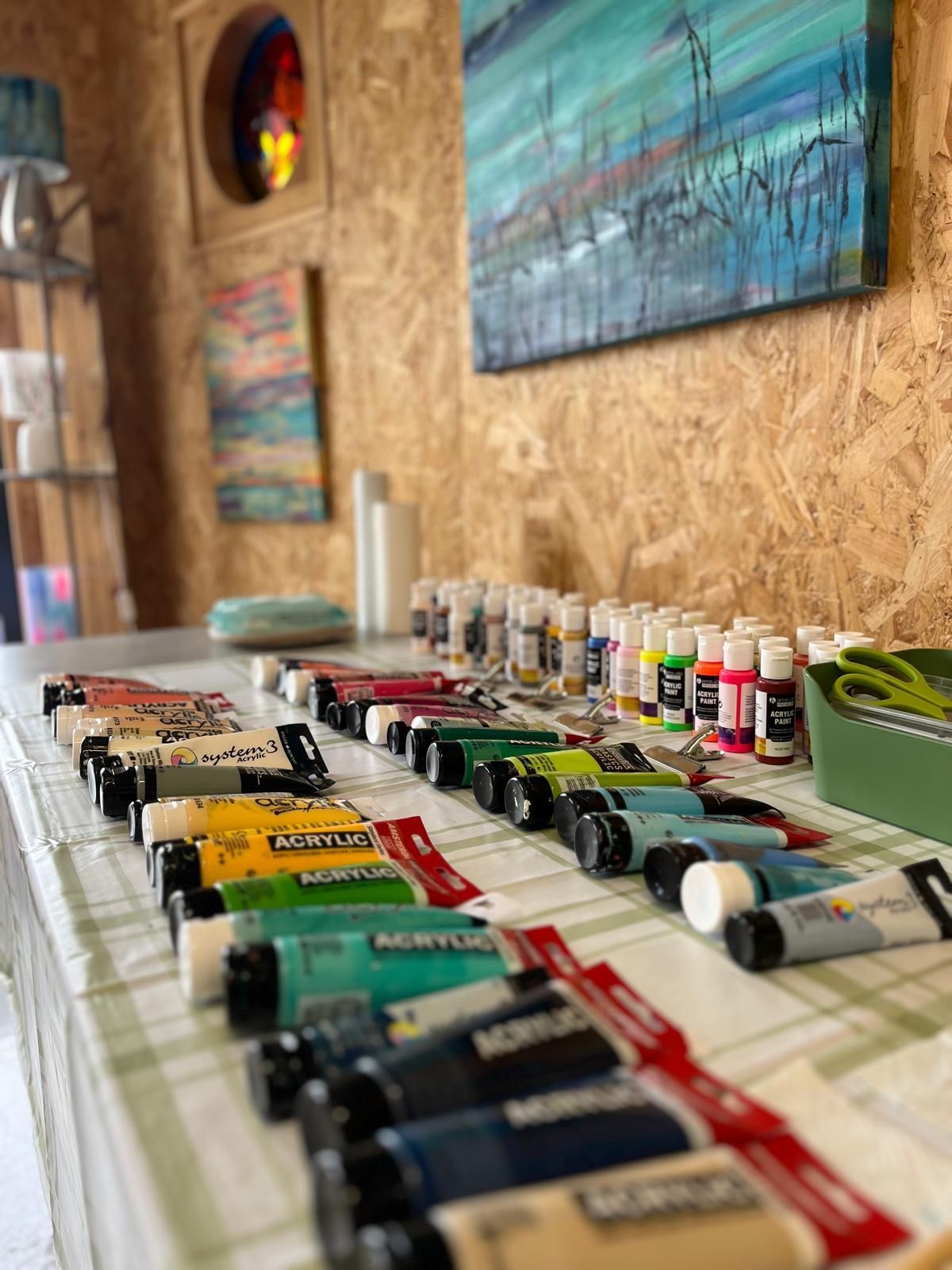 Tubes of acrylic paint on a table with paintings in the background in an art studio.