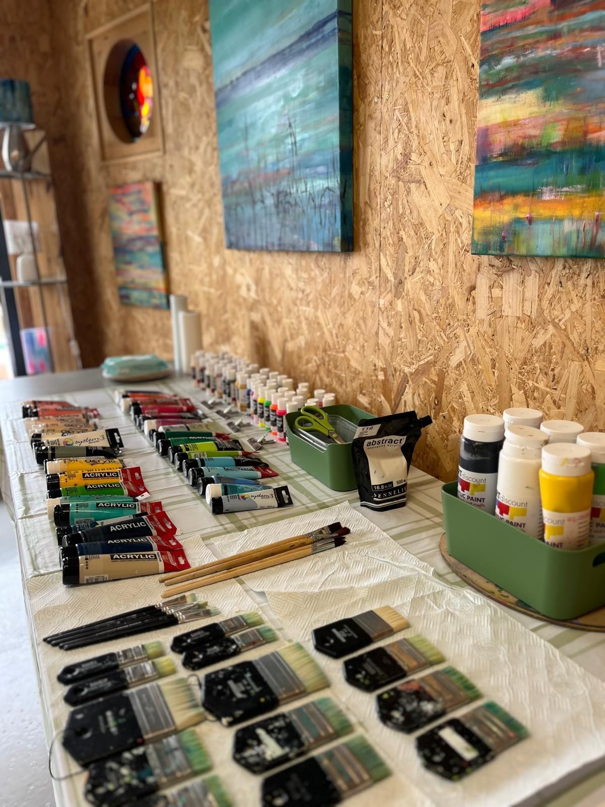 Art studio with paintings, paint brushes, and tubes of paint on a table. Wood panel walls.