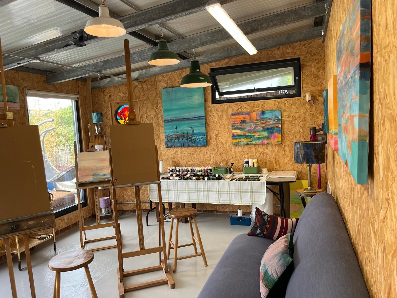 Art studio interior with easels, paintings, and a couch. Natural light, wood-paneled walls, and colorful art.