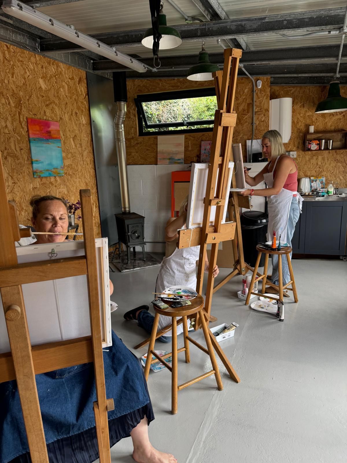 Three women painting in an art studio. One is seated, another is standing at an easel, and a third looks toward the camera.